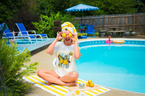 A woman sits cross-legged by a swimming pool wearing a white graphic t-shirt with a yellow lemon print, a yellow and white striped towel wrapped around her head, and yellow sunglasses. She is holding lemons in her hands, with a yellow and white striped towel on the pool deck in front of her.