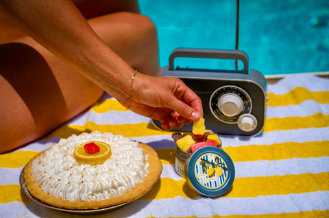 A lifestyle scene by a swimming pool showing a person's hand reaching for a small jar of fruit skewers on a yellow and white striped towel. In the foreground is a meringue-covered pie with a yellow lemon wheel garnish, and behind it is a retro-style portable radio.