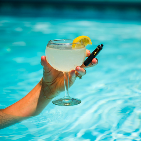 Hand emerging from a pool holding a lemon-garnished cocktail coupe and Appalachian Standards new Limoncello vape pen, poolside on a sunny day.