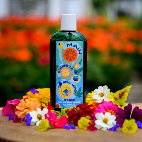 A bottle of Appalachian Standard's Main Squeeze Hemp Lotion on a piece of wood surrounded by flowers
