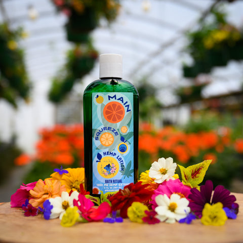 A bottle of Appalachian Standard's Main Squeeze Hemp Lotion on a piece of wood surrounded by flowers