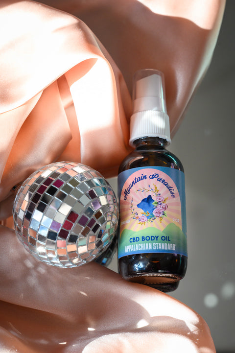 Appalachian Standard's Mountain Paradise CBD Body Oil with a pink satin sheet and shiny disco ball.