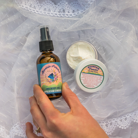 A hand reaching for an amber bottle with a blue/pink label that says "Mountain Paradise Body Oil" beside a open body cream container that says "Mountain Paradise" all on a white lace fabric