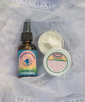 A hand reaching for an amber bottle with a blue/pink label that says "Mountain Paradise Body Oil" beside a open body cream container that says "Mountain Paradise" all on a white lace fabric