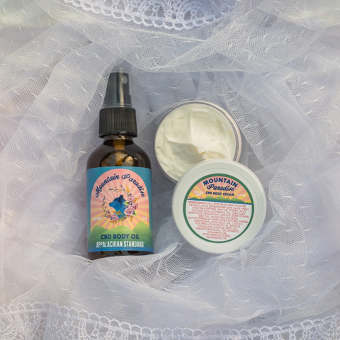 A hand reaching for an amber bottle with a blue/pink label that says "Mountain Paradise Body Oil" beside a open body cream container that says "Mountain Paradise" all on a white lace fabric