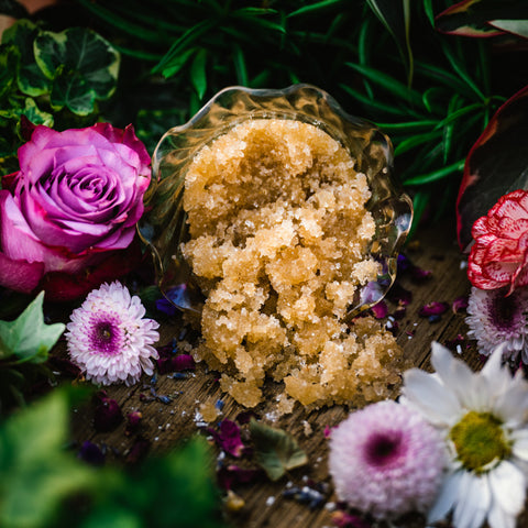 Sugar Scrub in a glass jar spilling on a table surrounded by herbs and flowers