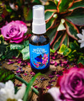 A 2 oz bottle of Appalachian Standard's For the Love of Flowers Body Oil with CBD on a wooden table surrounded by roses and lavender