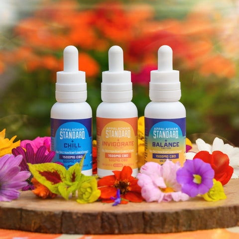 Three hemp oil tinctures with CBD, CBG, and CBN on top of a wooden platter surrounded by flowers by Appalachian Standard.
