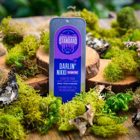 A tin of Appalachian Standard's Darlin' Nikki 4 a Funky Time Indica Flight standing upright on a bed of moss with wood slices in the background