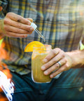 A man in a plaid tshirt and jeans dropping hemp tincture into a mason jar of orange chai.