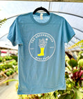 A close-up view of the same light blue Ross Farm Greenhouses t-shirt hanging from a metal pipe in a greenhouse structure. The shirt displays the circular logo with the yellow boot filled with illustrated flowers and plants.