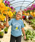 A woman with short gray hair and glasses smiling in a greenhouse filled with colorful flowers. She's wearing the blue Ross Farm Greenhouses t-shirt and black shorts while holding a bunch of yellow bougainvillea or similar flowering plants, surrounded by vibrant pink and yellow flowers on display.