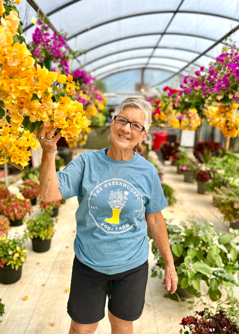 A woman with short gray hair and glasses smiling in a greenhouse filled with colorful flowers. She's wearing the blue Ross Farm Greenhouses t-shirt and black shorts while holding a bunch of yellow bougainvillea or similar flowering plants, surrounded by vibrant pink and yellow flowers on display.