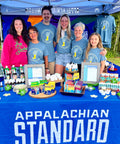 A group of six people of different ages standing behind a booth with "APPALACHIAN STANDARD" banner. They're all wearing the blue Ross Farm Greenhouses t-shirts (except one in pink), displaying various health and body care products on a blue-covered table at what appears to be a market or fair.