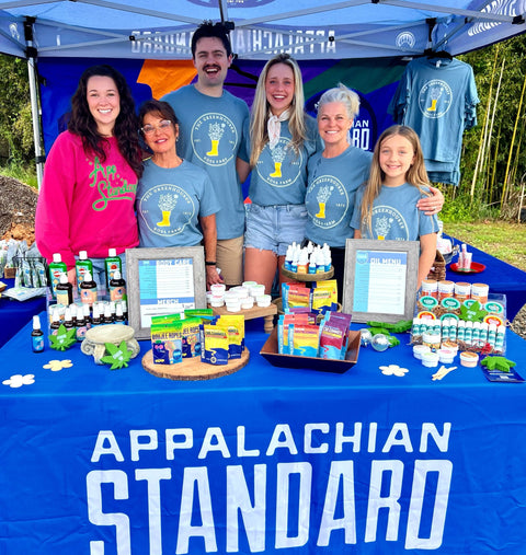 A group of six people of different ages standing behind a booth with "APPALACHIAN STANDARD" banner. They're all wearing the blue Ross Farm Greenhouses t-shirts (except one in pink), displaying various health and body care products on a blue-covered table at what appears to be a market or fair.