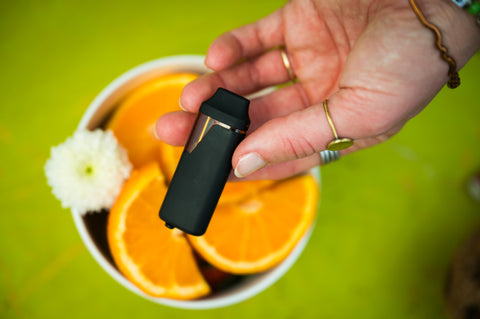 Hand holding a black vape device with a bowl of oranges and a white flower on a green background