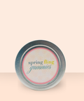 Top view of the Appalachian Standard Spring Fling Gummies tin lid on a soft peach background, featuring a round label with "spring fling gummies" in teal, gold, and script lettering on a daisy-patterned background with a coral border.