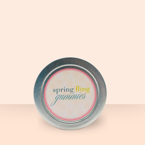 Top view of the Appalachian Standard Spring Fling Gummies tin lid on a soft peach background, featuring a round label with "spring fling gummies" in teal, gold, and script lettering on a daisy-patterned background with a coral border.