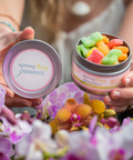 A woman's hands hold open the Appalachian Standard Spring Fling Gummies tin, filled with colorful sugar-coated gummies, alongside the tin lid, surrounded by fresh pink and orange orchid blooms.