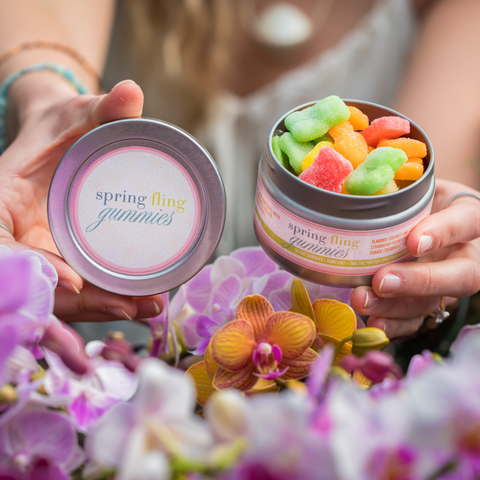 A woman's hands hold open the Appalachian Standard Spring Fling Gummies tin, filled with colorful sugar-coated gummies, alongside the tin lid, surrounded by fresh pink and orange orchid blooms.