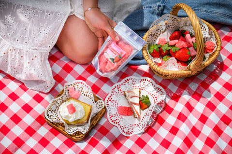 Overhead view of a picnic setup on a classic red and white checkered blanket. A hand holds a plastic package of pink strawberry-shaped gummies next to a wicker basket containing fresh strawberries and more pink gummies. Small decorative doilies hold desserts including pound cake topped with whipped cream and a strawberry with pink gummies as garnish.