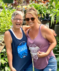 Two women standing together in a garden setting, wearing tank tops with visible text.