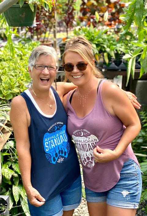 Two women standing together in a garden setting, wearing tank tops with visible text.