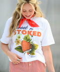 A person with long blonde hair looking down with a smile, wearing a white "Sweet n' Stoked" t-shirt featuring a detailed botanical illustration of a strawberry with leaves and flowers. The person has a red bandana tied around their neck, turquoise rings, and is wearing red and white striped shorts. The brand name "Appalachian Standard" appears at the bottom of the design.