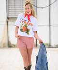 A full-body shot of someone wearing the same "Sweet n' Stoked" strawberry t-shirt with red and white striped shorts, brown boots, and a red bandana. They're smiling, looking to the side, and holding a denim jacket while standing against a white industrial backdrop. The outfit creates a cohesive red and white summer aesthetic.
