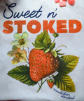 A close-up detail shot of the "Sweet n' Stoked" t-shirt graphic showing the botanical strawberry illustration against a white background with a red checkered border. The design features detailed strawberry fruit with green leaves and delicate strawberry blossoms. Some pink strawberry-shaped gummies are visible at the top of the image. The "Appalachian Standard" logo appears at the bottom of the design.