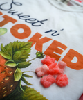 An extreme close-up of the "Sweet n' Stoked" t-shirt design focusing on the brand name in orange letters and part of the strawberry illustration. Several pink strawberry-shaped gummies are placed on the shirt, showing both the product and the matching graphic design theme.