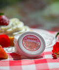 A small white container of Sweet n' Stoked CBD body cream (1.5 oz) on a red and white checkered tablecloth, surrounded by strawberries and red flowers. The product label indicates it contains 188 mg of CBD and lists various natural ingredients.