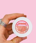 A hand holding the Sweet n' Stoked CBD body cream against a pink background, providing scale and showing the same silver ring with turquoise stone. The product's ingredient list is visible on the label.