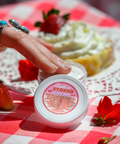 A close-up view of someone's hand with turquoise jewelry holding or pointing to the same Sweet n' Stoked CBD body cream. The product is placed on what appears to be a decorative plate with a dessert topped with whipped cream and a strawberry in the background, still on the red and white checkered tablecloth.