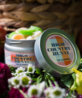 Open tin of Appalachian Standard High Country Hunny THC Gummies showing an assortment of gourmet flavored gummies in orange, green, and white, styled with red apples, daisies, and yellow flowers against a woven basket background.