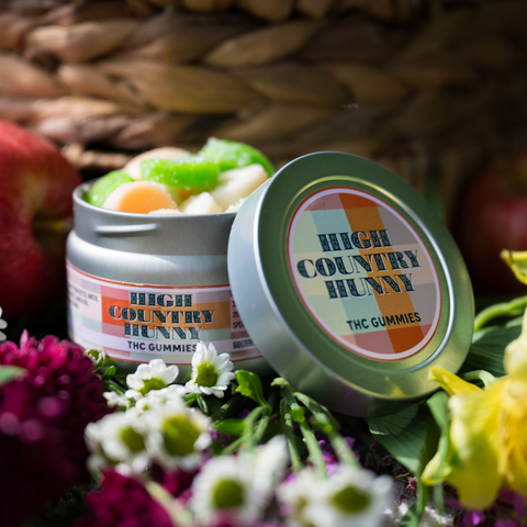 Open tin of Appalachian Standard High Country Hunny THC Gummies showing an assortment of gourmet flavored gummies in orange, green, and white, styled with red apples, daisies, and yellow flowers against a woven basket background.