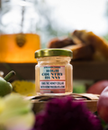 

Jar of Appalachian Standard High Country Hunny 15mg THC-infused honey, 2oz, collaboration with HoneyBeeBliss.com, displayed on a wooden table with fresh apples, a pear, purple flowers, and a blurred honey dispenser and baked goods in the background.