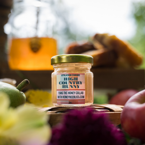 

Jar of Appalachian Standard High Country Hunny 15mg THC-infused honey, 2oz, collaboration with HoneyBeeBliss.com, displayed on a wooden table with fresh apples, a pear, purple flowers, and a blurred honey dispenser and baked goods in the background.