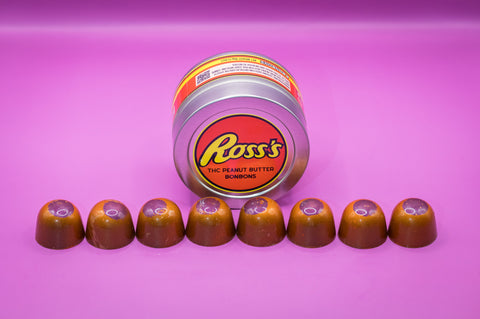 Appalachian Standard's limited edition Ross's THC Peanut Butter BonBons tin lying on its side with eight airbrushed orange bonbons lined up in front, on a purple background