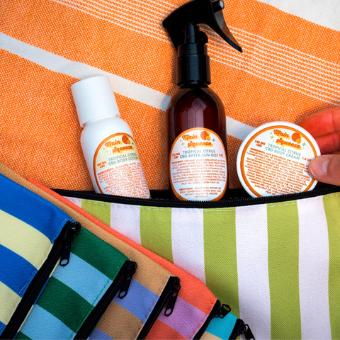Appalachian Standard Tropical Citrus Main Squeeze CBD skincare trio — body lotion, after-sun mist spray, and body cream — displayed in one of the 6 colorful striped travel pouches they come with, on an orange beach towel.