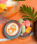 Appalachian Standard Acapulco Gold Gummies tin open on an orange beach towel, filled with colorful assorted CBD gummies, surrounded by a tropical cocktail, palm leaf, and coconut.