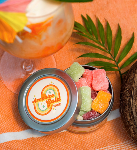 Appalachian Standard Acapulco Gold Gummies tin open on an orange beach towel, filled with colorful assorted CBD gummies, surrounded by a tropical cocktail, palm leaf, and coconut.