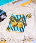 Cream lemon tee shirt on a striped yellow and white towel with a grey radio by a blue pool