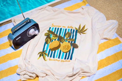 Cream lemon tee shirt on a striped yellow and white towel with a grey radio by a blue pool
