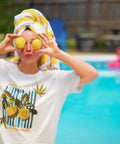 Woman in front of a blue pool with floats in it holding two yellow lemons over eyes with yellow and white towel in hair, wearing a cream lemon t-shirt