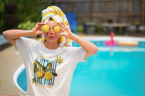 Woman in front of a blue pool with floats in it holding two yellow lemons over eyes with yellow and white towel in hair, wearing a cream lemon t-shirt