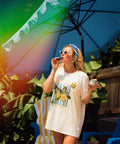 Woman standing under a blue umbrella with blue chairs behind while wearing a cream t-shirt with lemons on it