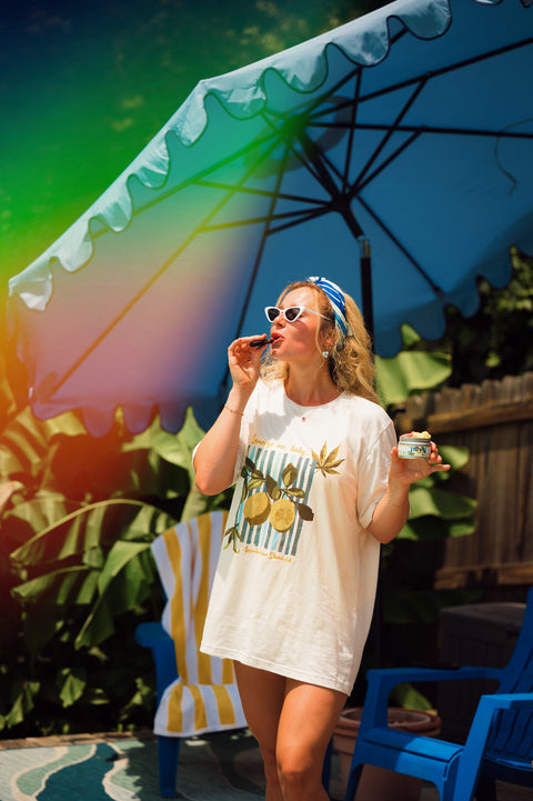 Woman standing under a blue umbrella with blue chairs behind while wearing a cream t-shirt with lemons on it