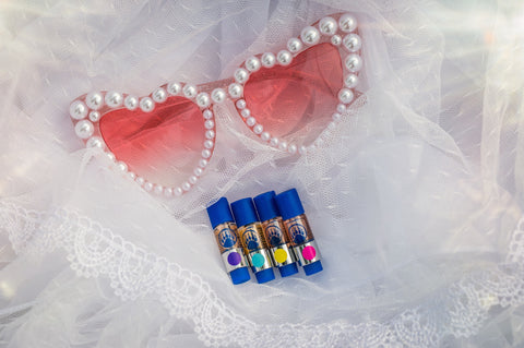 Pearl-embellished heart sunglasses with four colorful CBD oil vape cartridges in blue cases on white tulle fabric