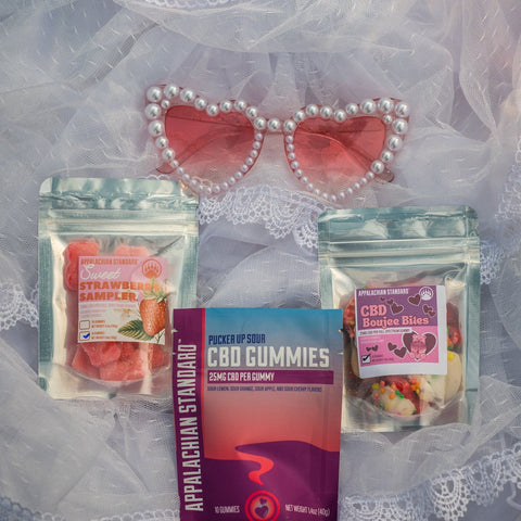 Pearl heart sunglasses displayed with Appalachian Standard CBD products including Pucker Up Sour gummies, Sweet Strawberry Sampler, and CBD Boujee Bites on lace fabric